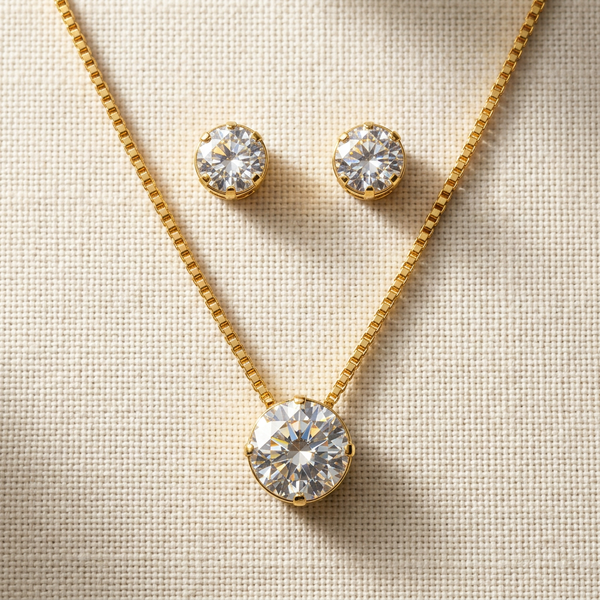 Conjunto de colar e brincos ponto de luz dourado, com pedra redonda de zircônia, apresentado em uma caixa de presente branca sobre um fundo de tecido neutro.
