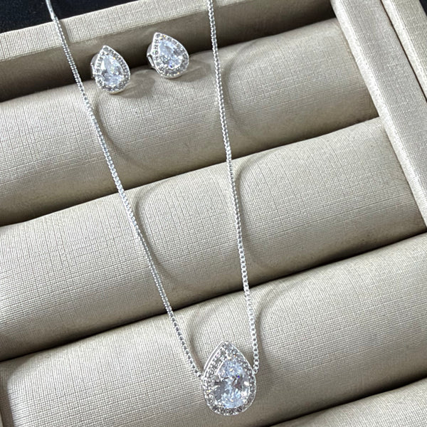 Conjunto de colar e brincos prateados com pingente e brincos em formato de gota, com uma pedra cristal central e um halo de pedras menores, sobre um mostruário bege.