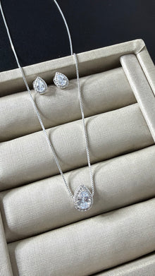Conjunto de colar e brincos prateados com pingente e brincos em formato de gota, com uma pedra cristal central e um halo de pedras menores, sobre um mostruário bege.