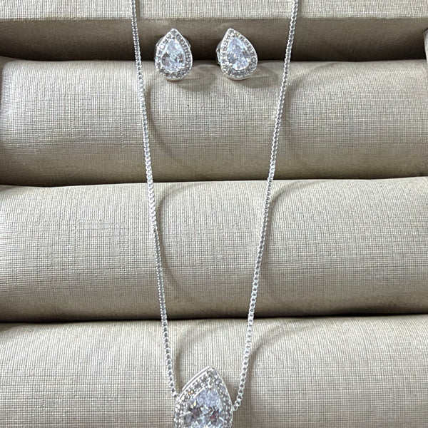 Conjunto de colar e brincos prateados com pingente e brincos em formato de gota, com uma pedra cristal central e um halo de pedras menores, sobre um mostruário bege.