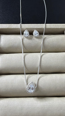 Conjunto de colar e brincos prateados com pingente e brincos em formato de gota, com uma pedra cristal central e um halo de pedras menores, sobre um mostruário bege.