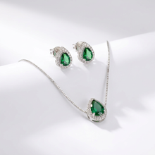 Conjunto de colar e brincos prateados com pingentes em formato de gota com pedra verde esmeralda central e contorno de zircônias, exibido em um estojo bege.
