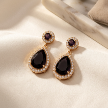 Brinco gota violeta dourado com cristais em fundo espelhado.