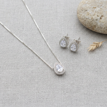 Conjunto de colar e brincos prateados com pingente e brincos em formato de gota, com uma pedra cristal central e um halo de pedras menores, sobre um mostruário bege.