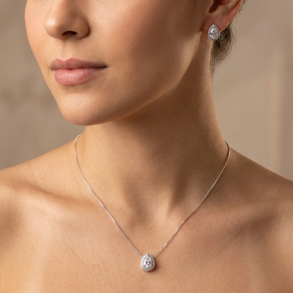 Conjunto de colar e brincos prateados com pingente e brincos em formato de gota, com uma pedra cristal central e um halo de pedras menores, sobre um mostruário bege.