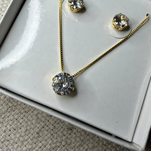 Conjunto de colar e brincos ponto de luz dourado, com pedra redonda de zircônia, apresentado em uma caixa de presente branca sobre um fundo de tecido neutro.