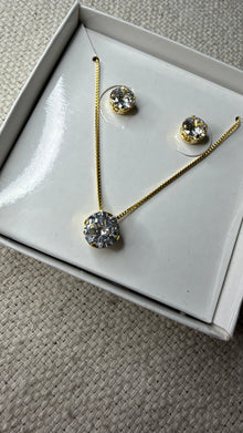 Conjunto de colar e brincos ponto de luz dourado, com pedra redonda de zircônia, apresentado em uma caixa de presente branca sobre um fundo de tecido neutro.