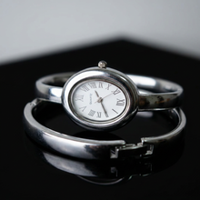 Relógio bracelete oval prata em aço inox com mostrador analógico e algarismos romanos.