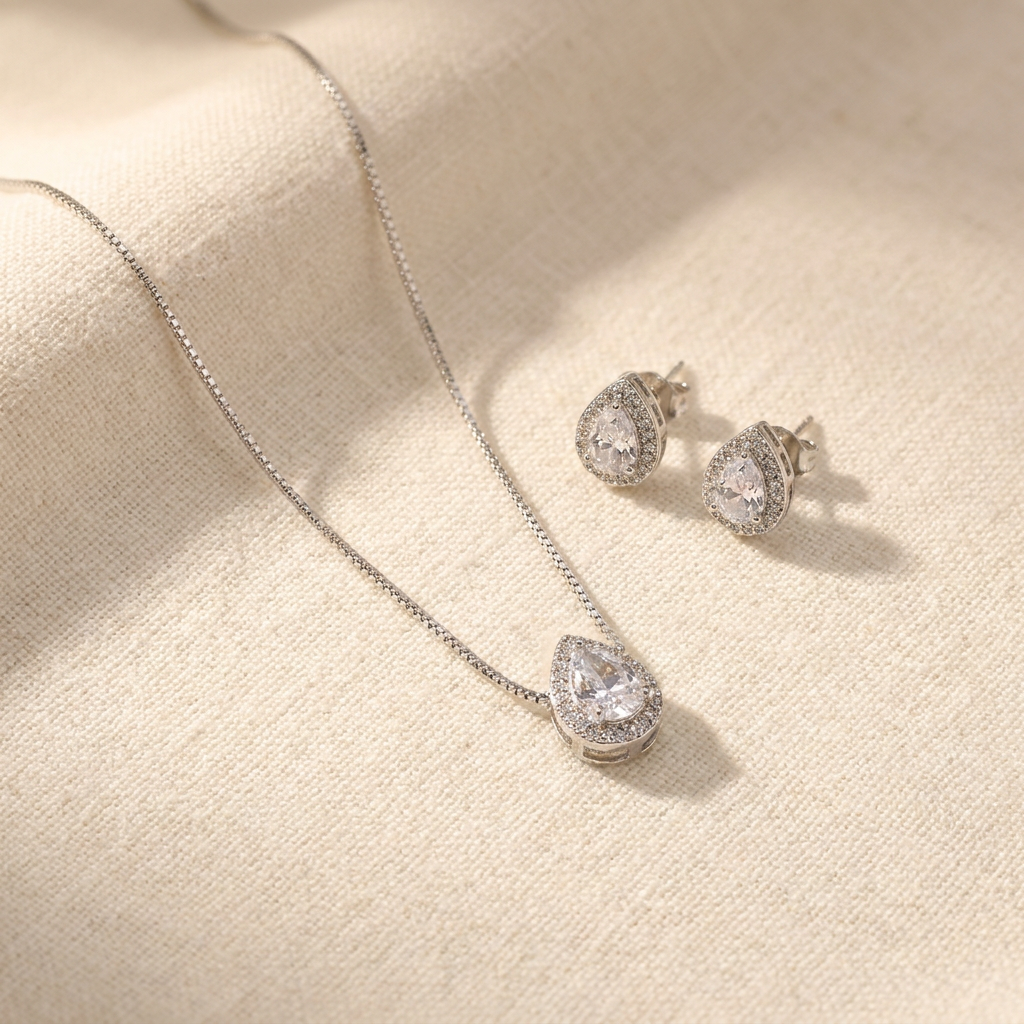 Conjunto de colar e brincos prateados com pingente e brincos em formato de gota, com uma pedra cristal central e um halo de pedras menores, sobre um mostruário bege.
