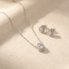 Conjunto de colar e brincos prateados com pingente e brincos em formato de gota, com uma pedra cristal central e um halo de pedras menores, sobre um mostruário bege.