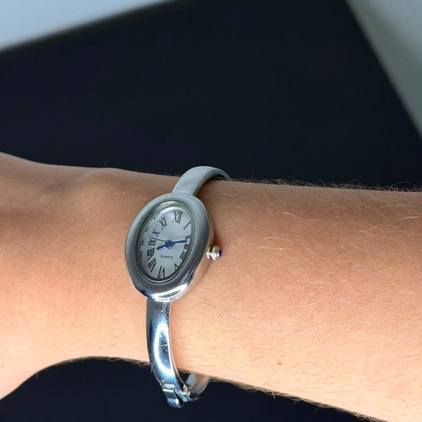 Relógio bracelete oval prata em aço inox com mostrador analógico e algarismos romanos.