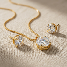 Conjunto de colar e brincos ponto de luz dourado, com pedra redonda de zircônia, apresentado em uma caixa de presente branca sobre um fundo de tecido neutro.