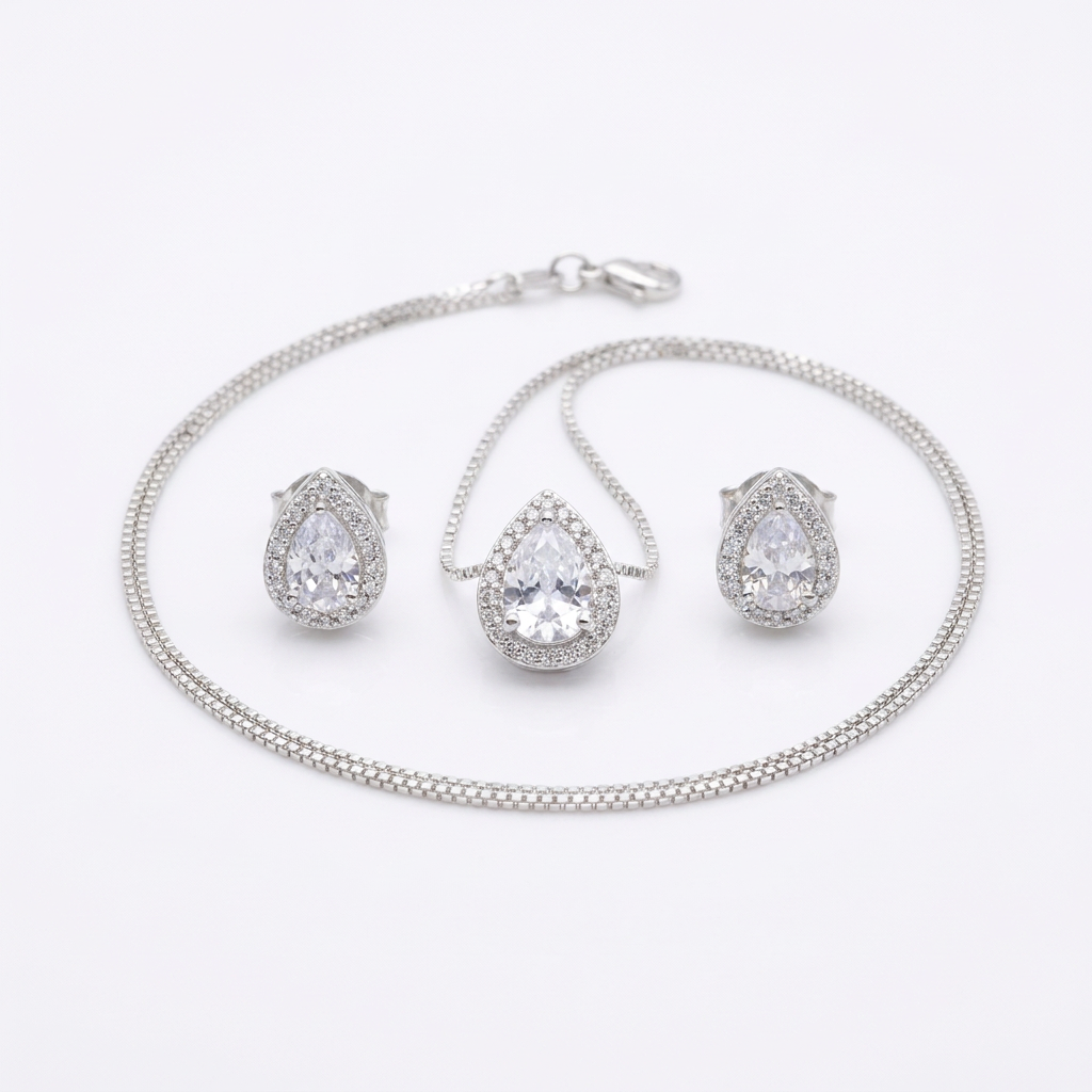 Conjunto de colar e brincos prateados com pingente e brincos em formato de gota, com uma pedra cristal central e um halo de pedras menores, sobre um mostruário bege.