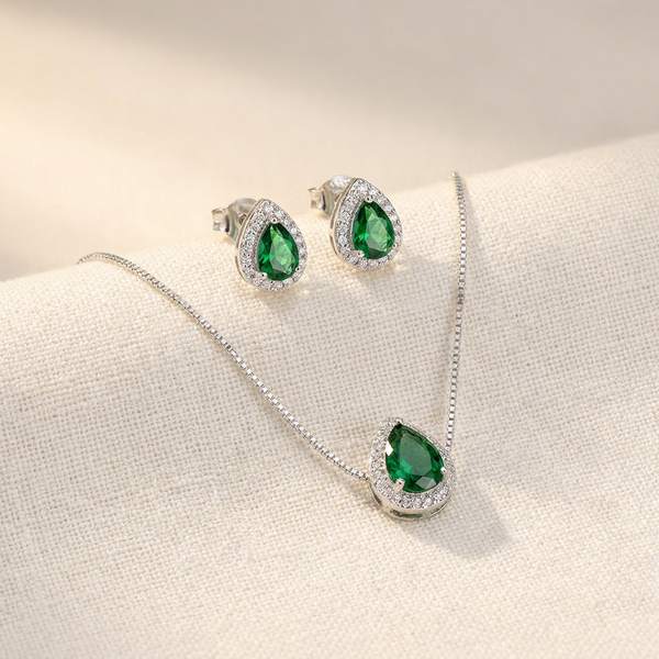 Conjunto de colar e brincos prateados com pingentes em formato de gota com pedra verde esmeralda central e contorno de zircônias, exibido em um estojo bege.