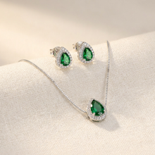 Conjunto de colar e brincos prateados com pingentes em formato de gota com pedra verde esmeralda central e contorno de zircônias, exibido em um estojo bege.