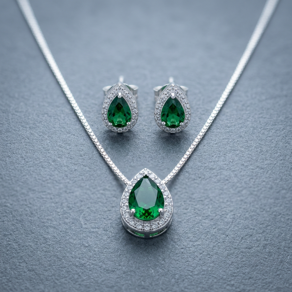 Conjunto de colar e brincos prateados com pingentes em formato de gota com pedra verde esmeralda central e contorno de zircônias, exibido em um estojo bege.
