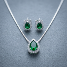 Conjunto de colar e brincos prateados com pingentes em formato de gota com pedra verde esmeralda central e contorno de zircônias, exibido em um estojo bege.