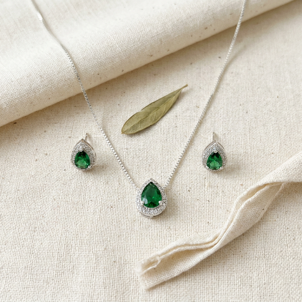 Conjunto de colar e brincos prateados com pingentes em formato de gota com pedra verde esmeralda central e contorno de zircônias, exibido em um estojo bege.